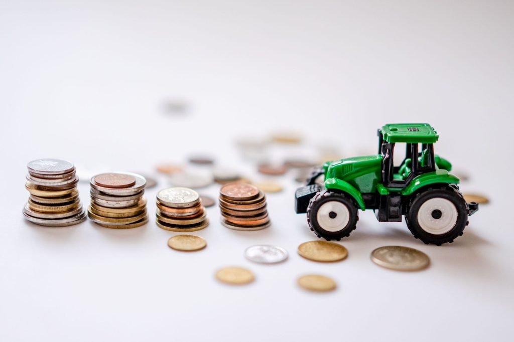 Safra 2026/27 começa com R$ 65,7 bilhões em crédito rural Miniatura de trator verde ao lado de pilhas de moedas sobre uma superfície clara.