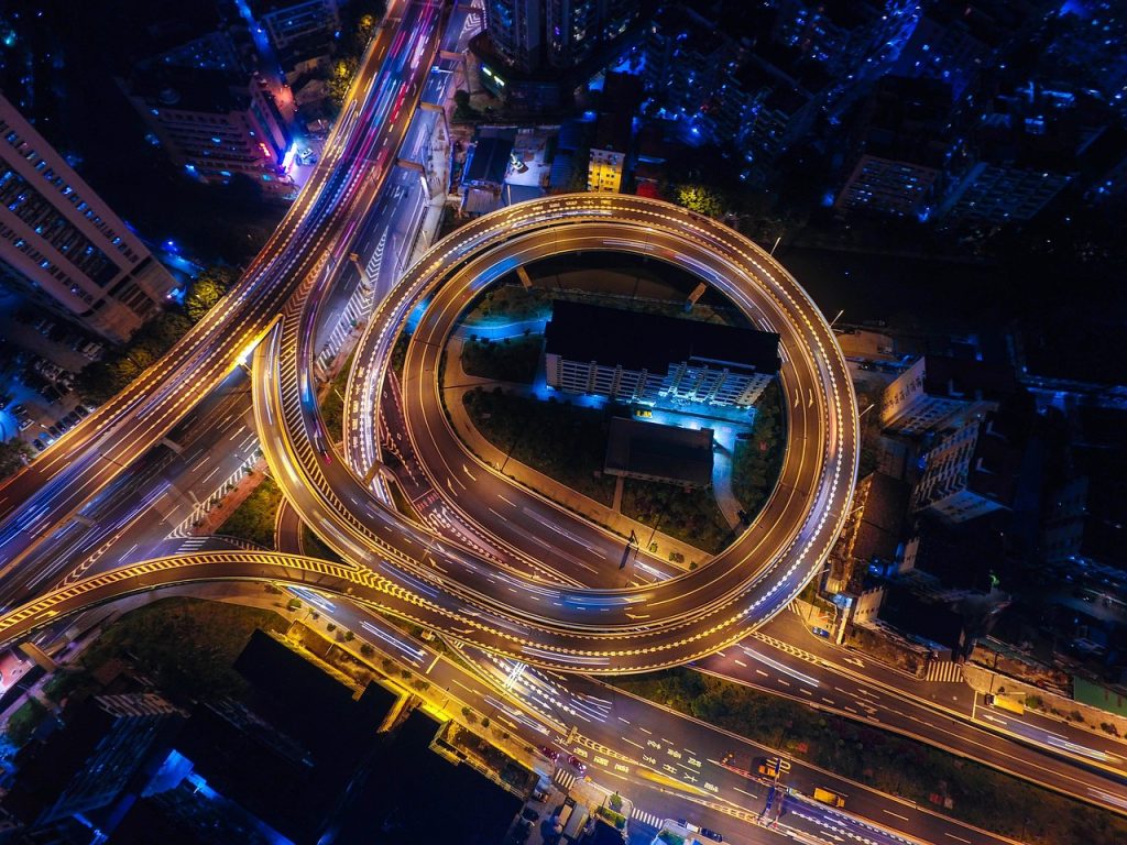 Vista aérea noturna de uma cidade, com ruas, cruzamentos e vias iluminadas pelo trânsito.